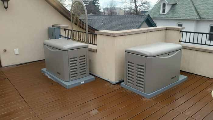 Two gray generators on a wooden deck next to a building with a railing, other buildings are in the background.