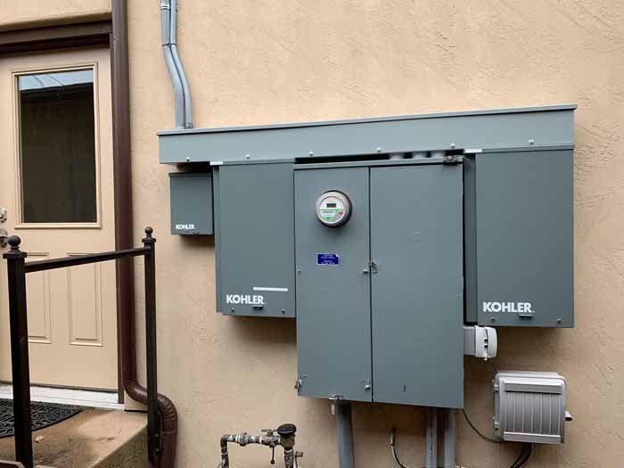 Gray electrical box mounted on a beige building exterior with a door and railing visible.