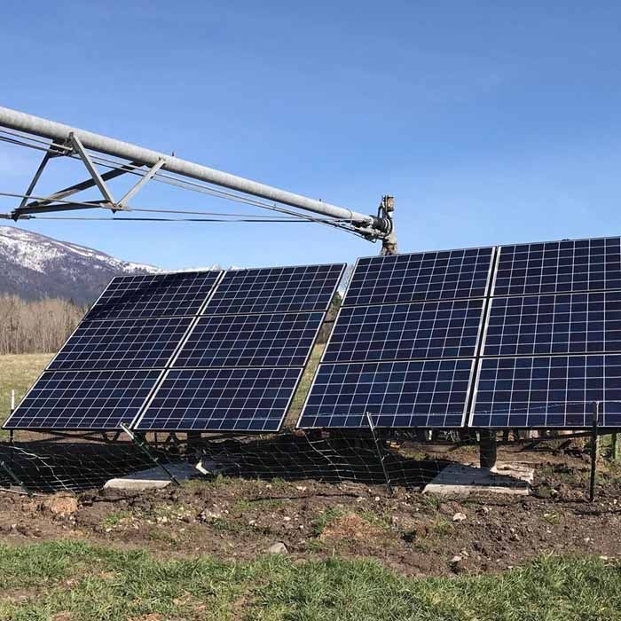 Solar panels in a field, with irrigation system overhead and mountains in the background.
