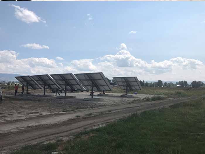 Solar panels installed outdoors, angled toward the sky, with people nearby. Cloudy blue sky and dirt road in the foreground.