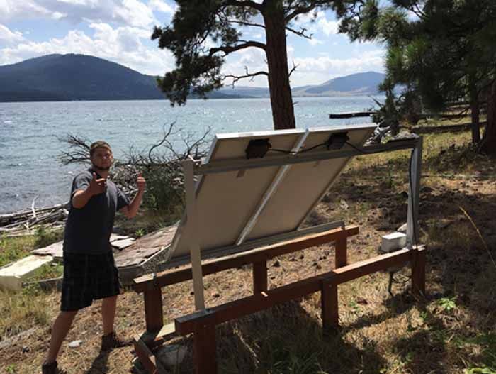 Man stands near a solar panel set near a lake and mountain range. He gives a thumbs up gesture.