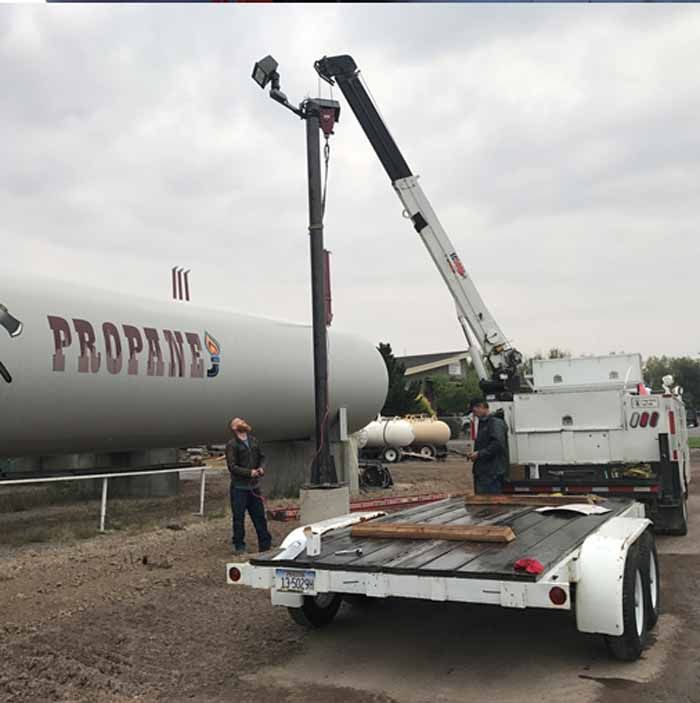 Crane working on a light pole beside a large propane tank. Two workers on site.