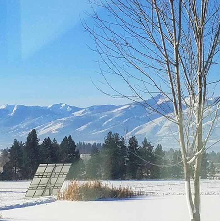 Snowy landscape with solar panels, trees, and mountains under a blue sky.