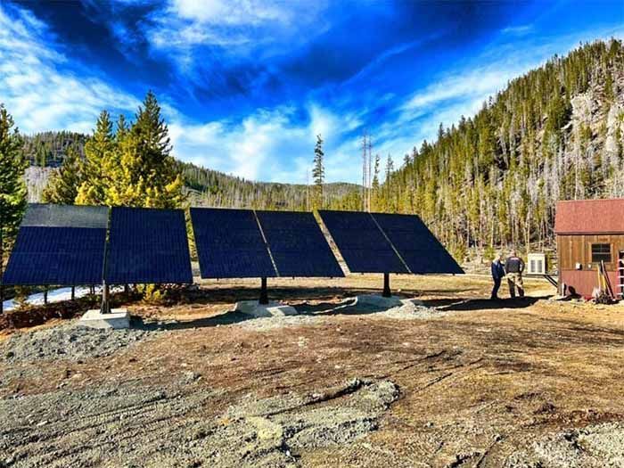 Solar panels in a mountain setting next to a small building, under a cloudy blue sky. Two people stand near the panels.