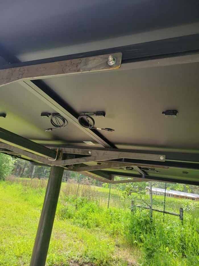 Underside view of a black metal structure, possibly a camper top, with supports and wiring, set outdoors with green grass.