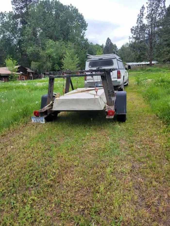 Trailer hitched to a white van on a grassy path, with trees and a building in the background.