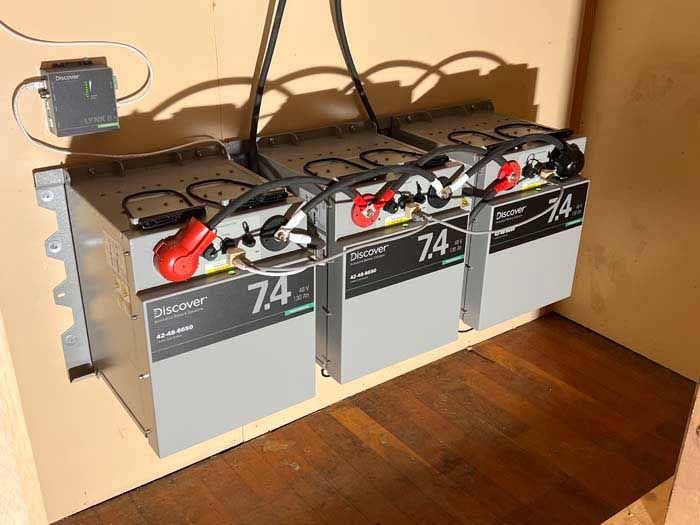 Three gray battery units with black cables and red/black terminals, mounted on a wooden surface.