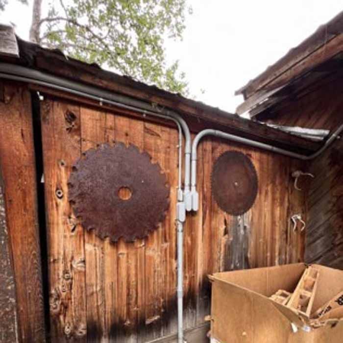 Wooden shed exterior with large rusty saw blades mounted, electrical conduit, and cardboard box.