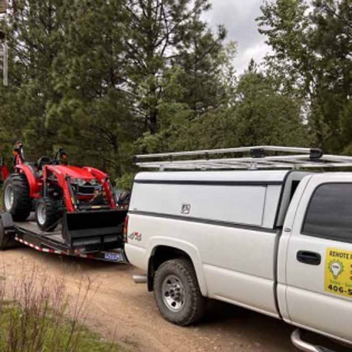 White truck towing a trailer with a red tractor on a dirt road in a wooded area.