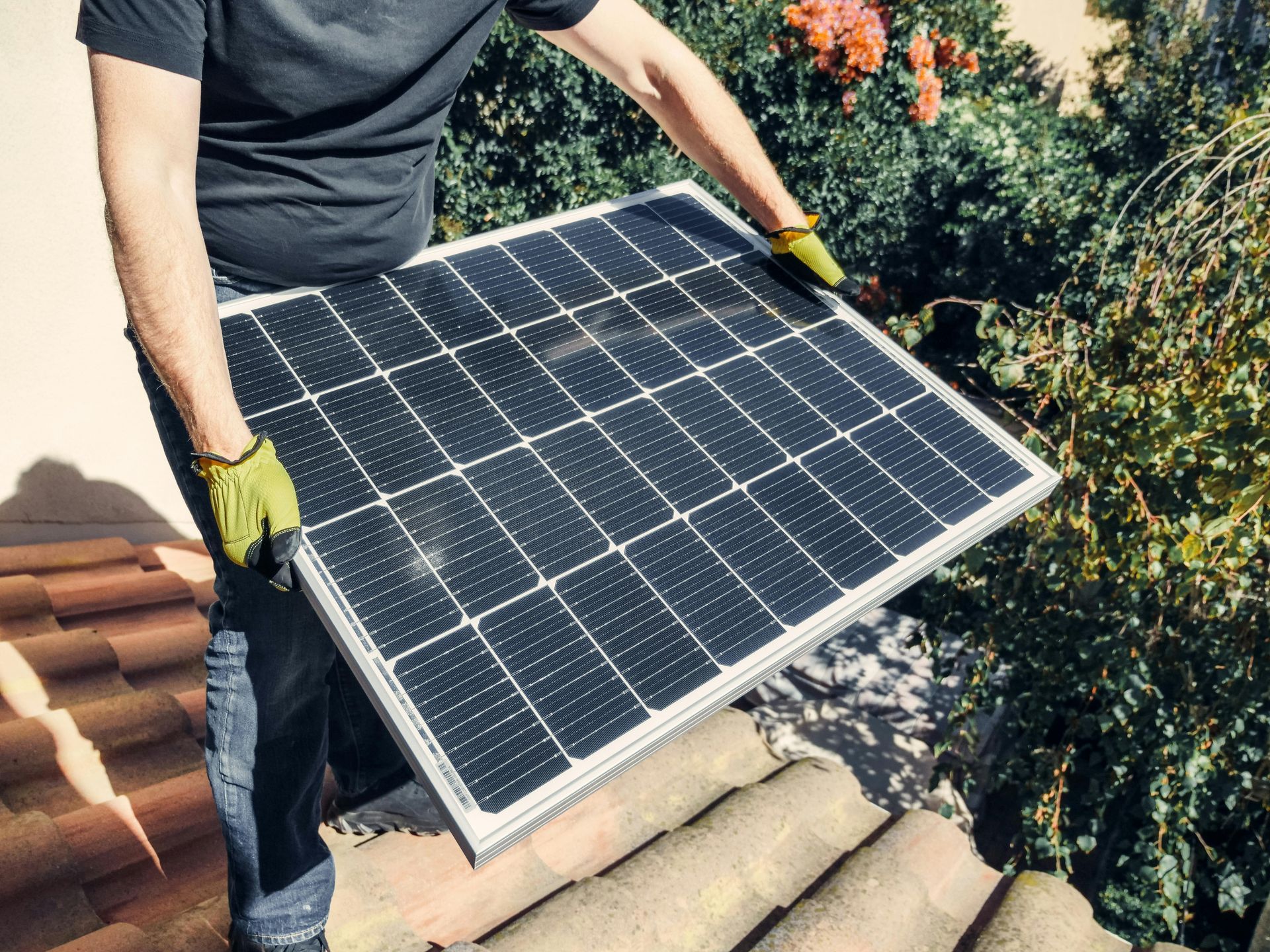 Person holding a solar panel on a tiled roof. Green gloves.