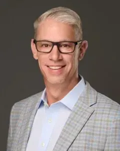 Man with glasses smiles, wearing a light blue shirt and plaid jacket.