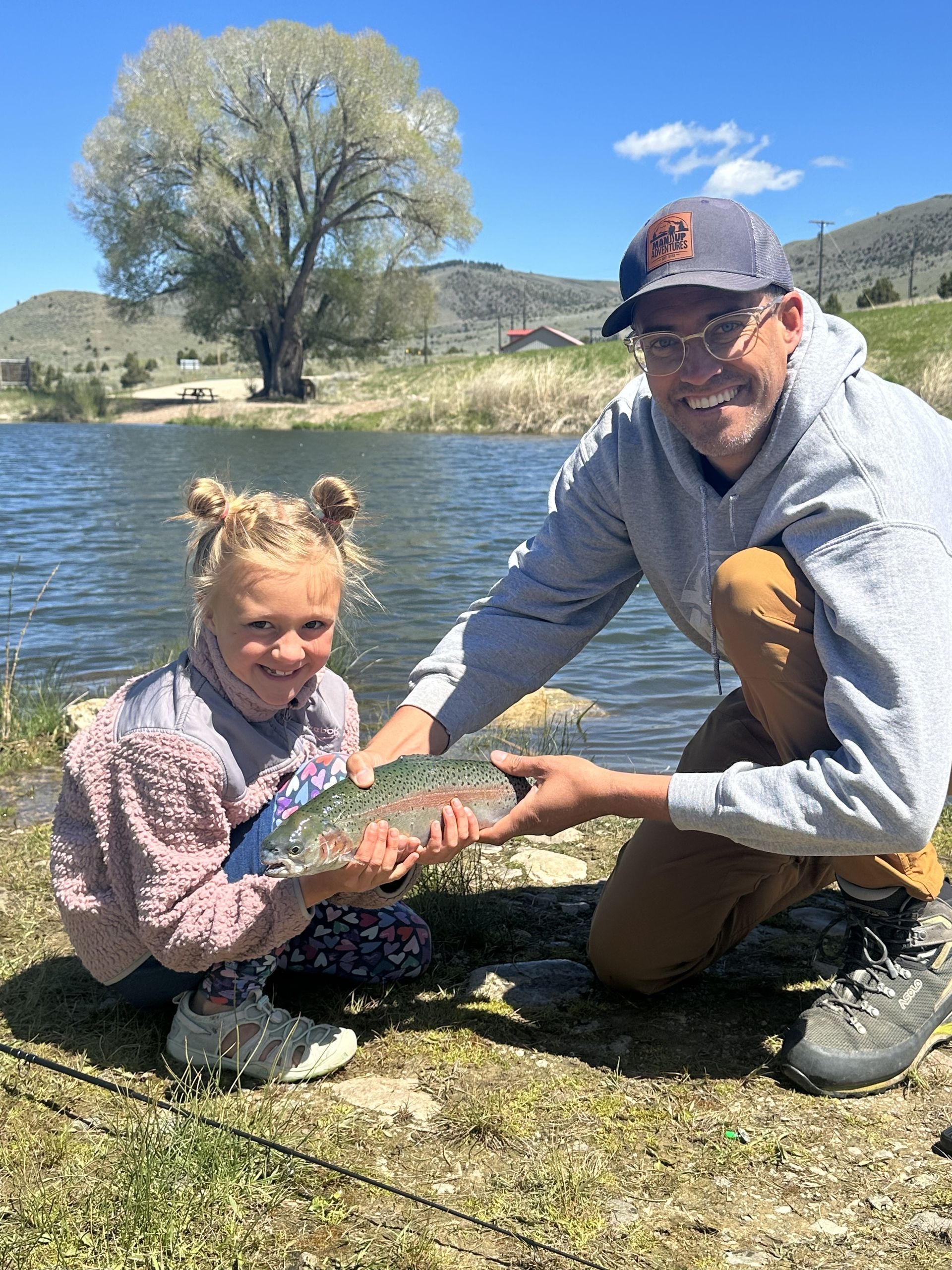 A man and a little girl are kneeling next to a lake holding a fish.