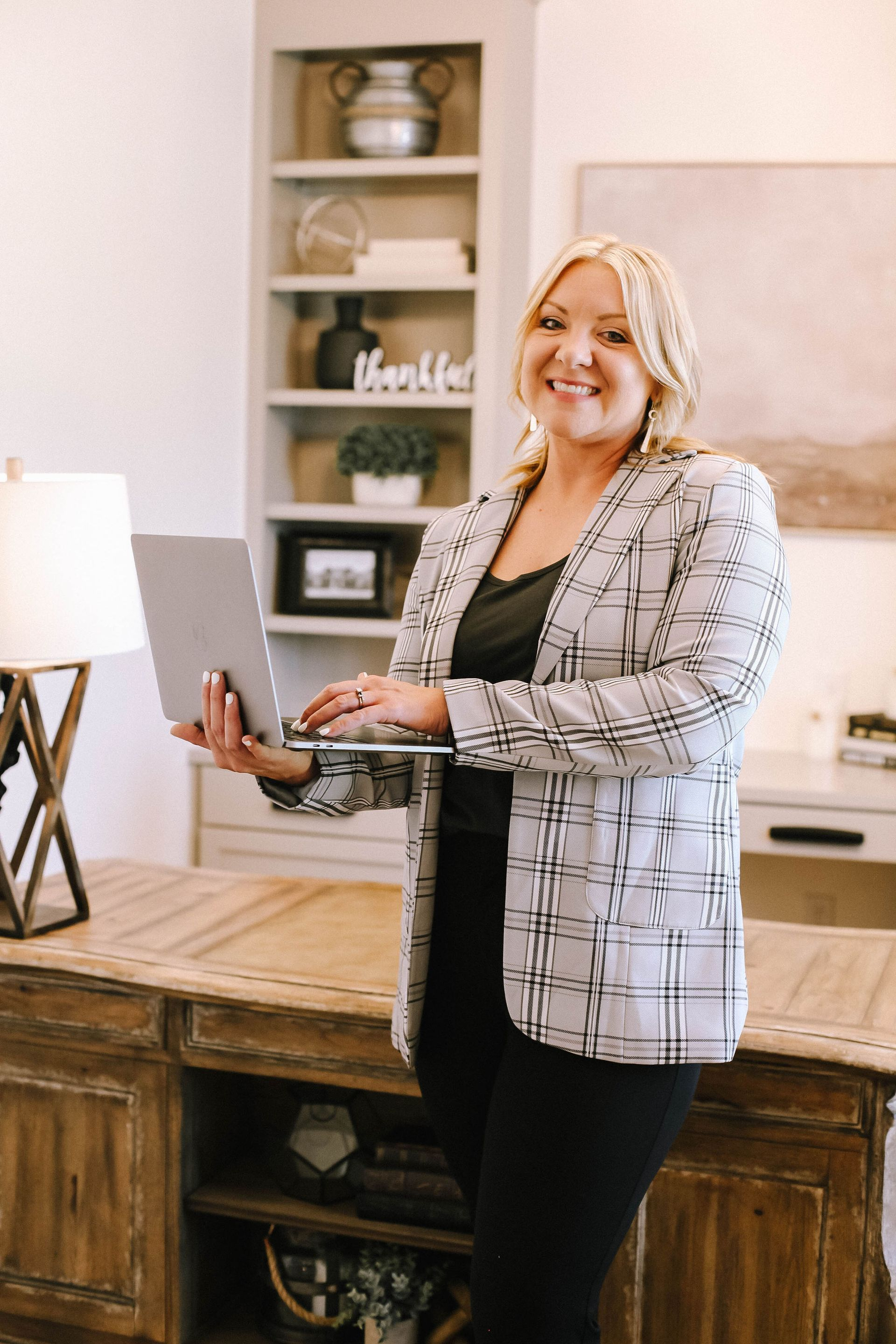 Woman in plaid blazer holds laptop, smiles in home office setting.