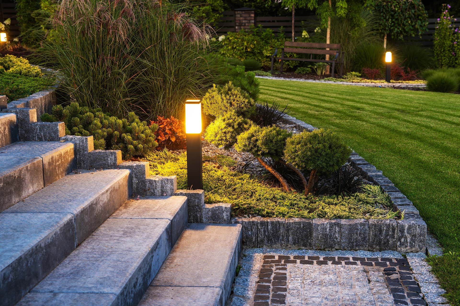 Stone steps lead up to a garden with illuminated path lights, lush greenery, and manicured lawn.