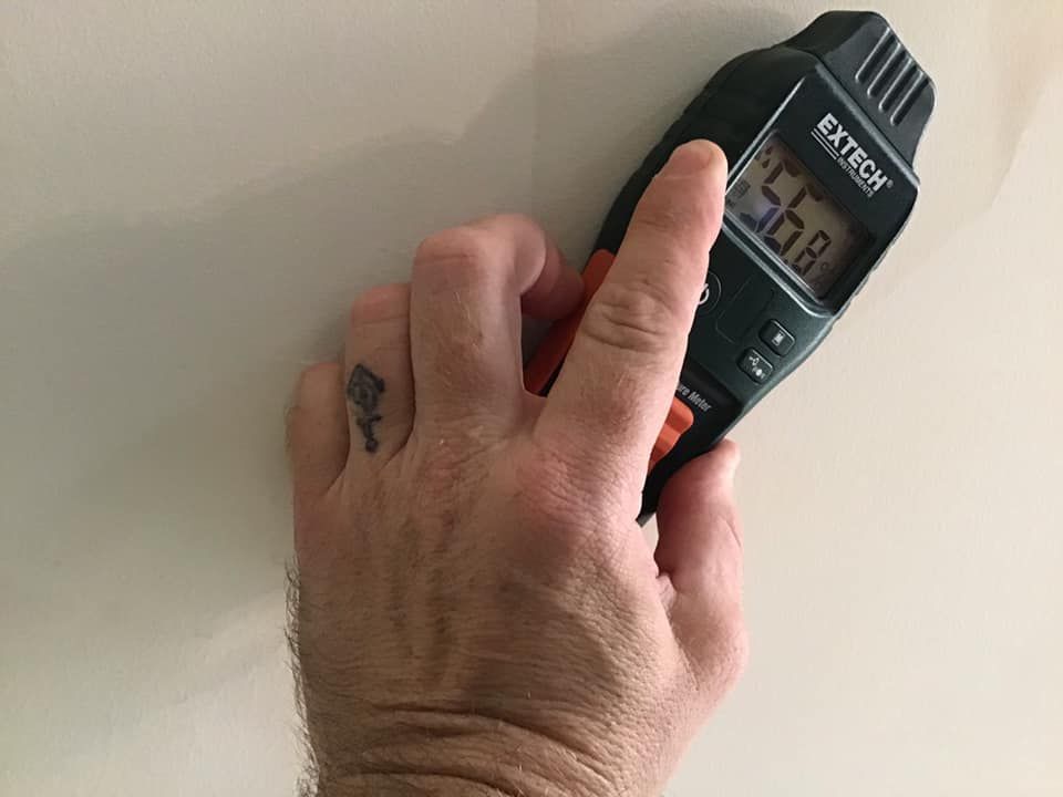 Hand holding a moisture meter against a cracked ceiling near a window frame. The meter reads 999.