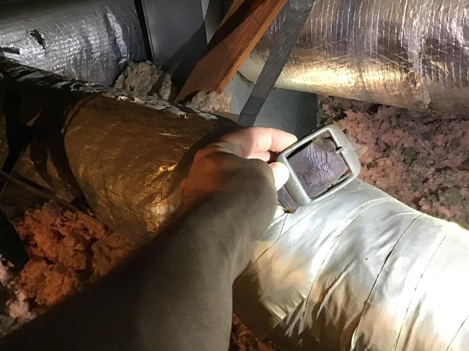 Hand holding a flashlight illuminates ductwork in an attic, with insulation visible.