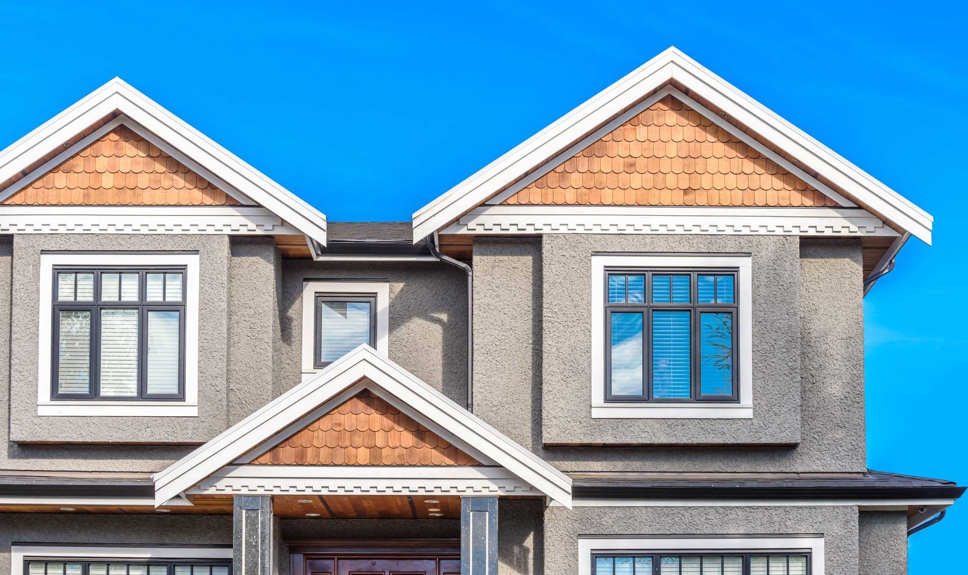 Two-story house with gray stucco, dark windows, and wood-paneled gables against a blue sky.