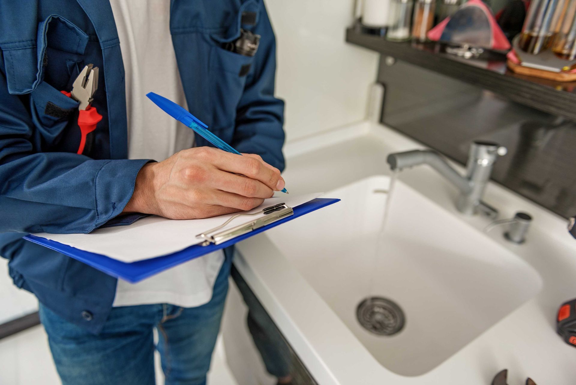 Plumber writing on a clipboard by a sink, wearing a blue uniform with tools.
