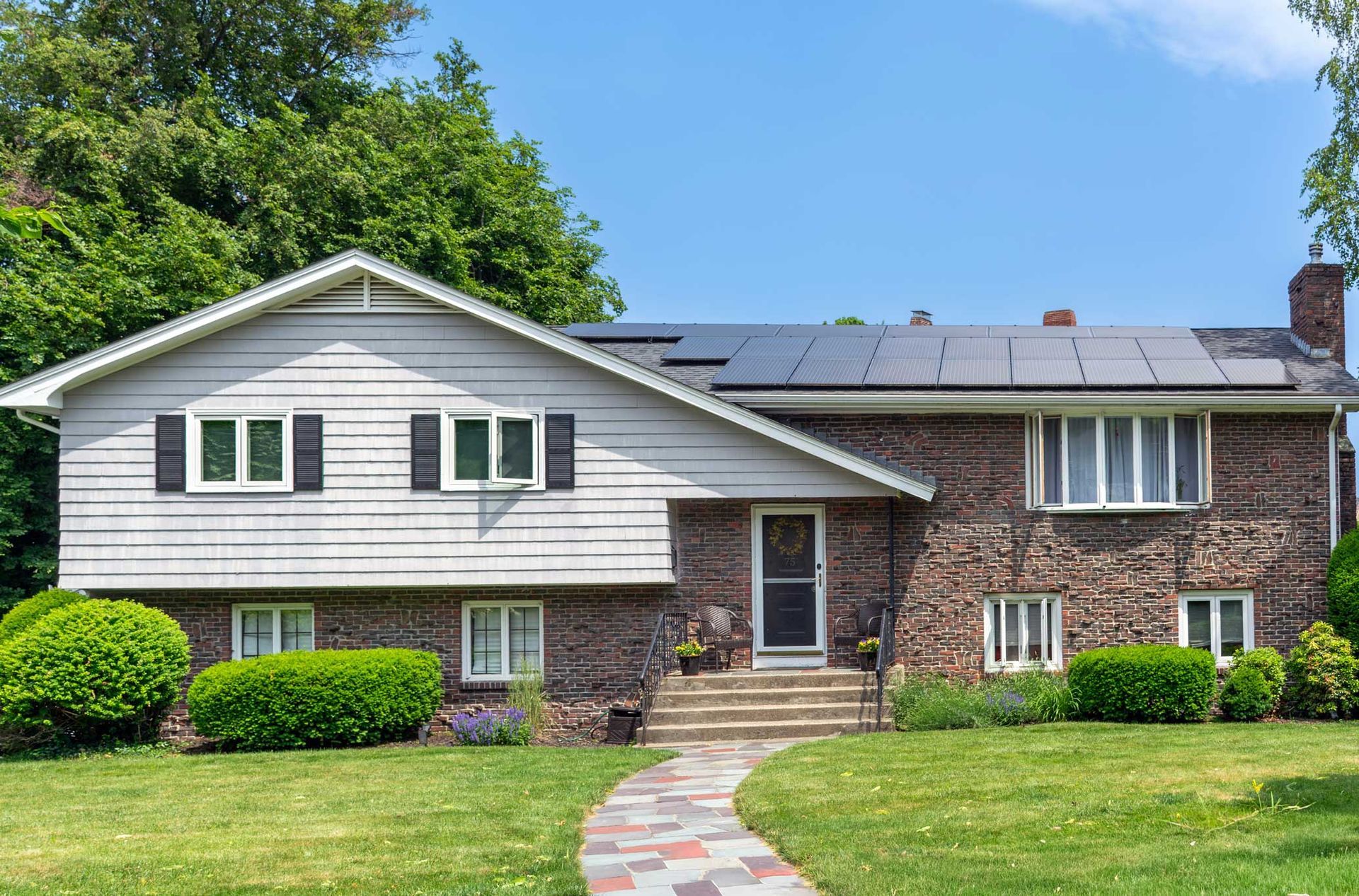 Two-story house with brick and siding, solar panels, and a stone walkway on a sunny day.