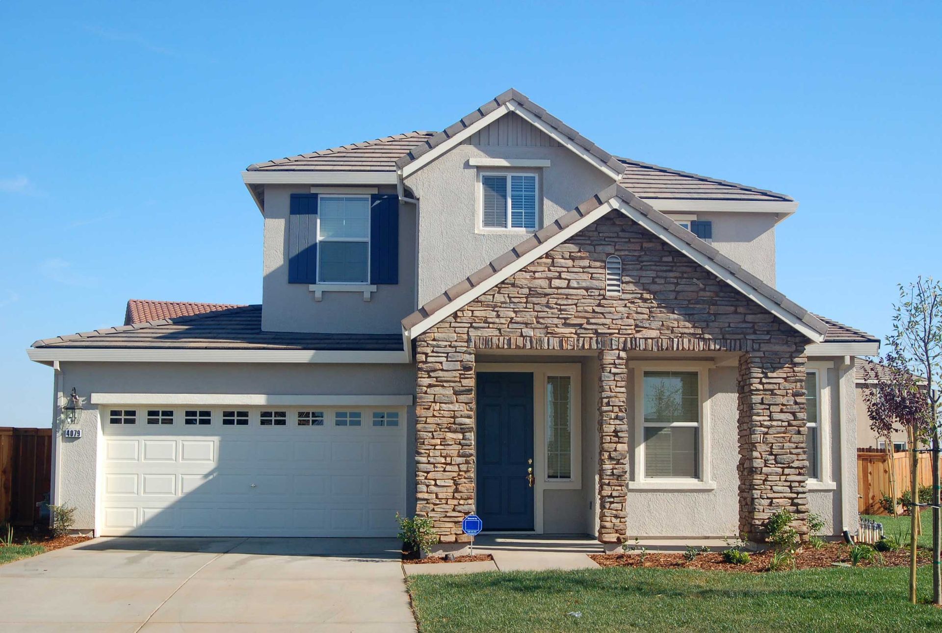 Two-story house with stone facade above front door, light gray stucco walls, blue shutters, and white garage door.