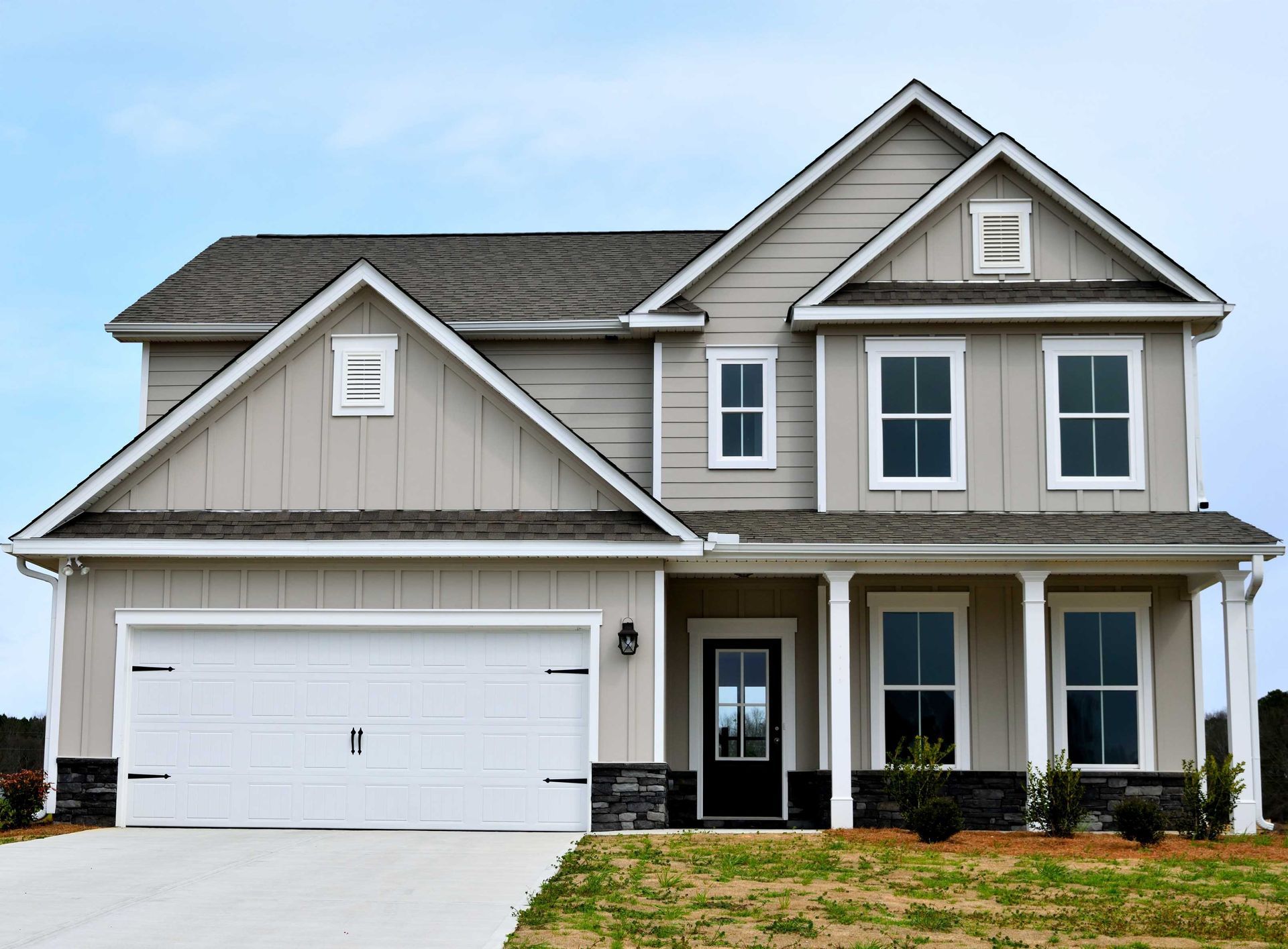 Two-story beige house with white trim, dark roof, front door, garage, and windows against a blue sky.
