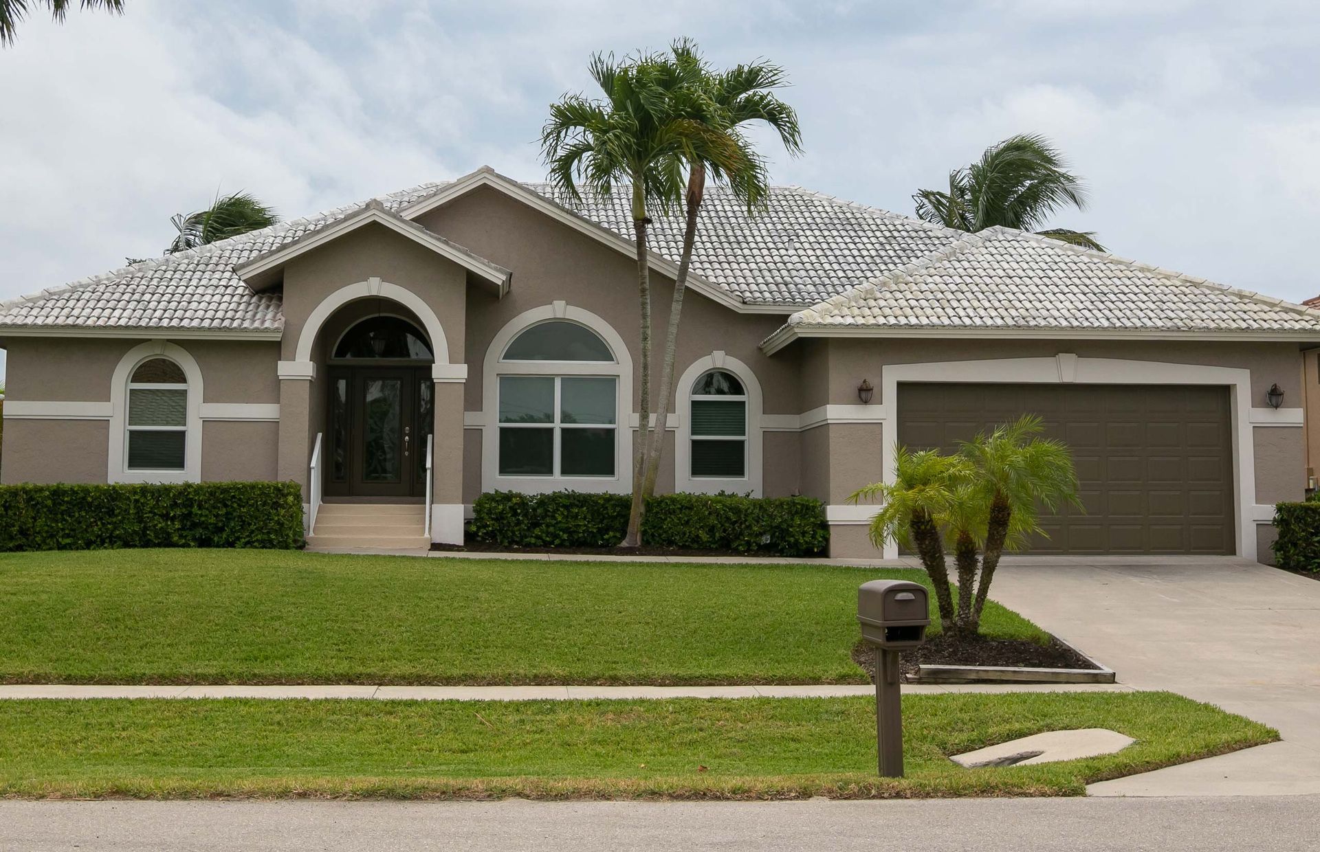 Tan house with a light gray tile roof, green lawn, palm trees, and brown garage door.