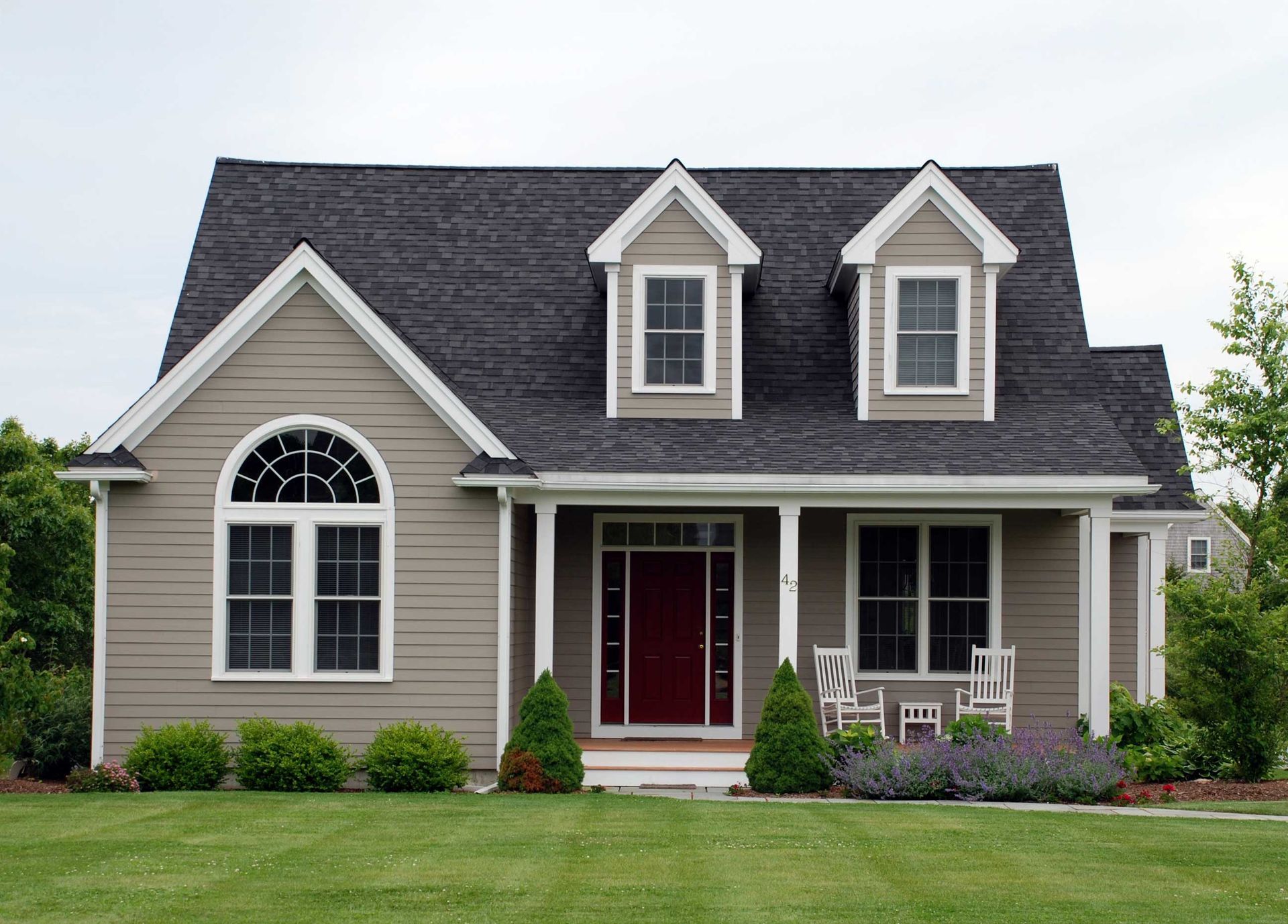 Tan house with dark gray roof, red door, dormer windows, and porch, green lawn.
