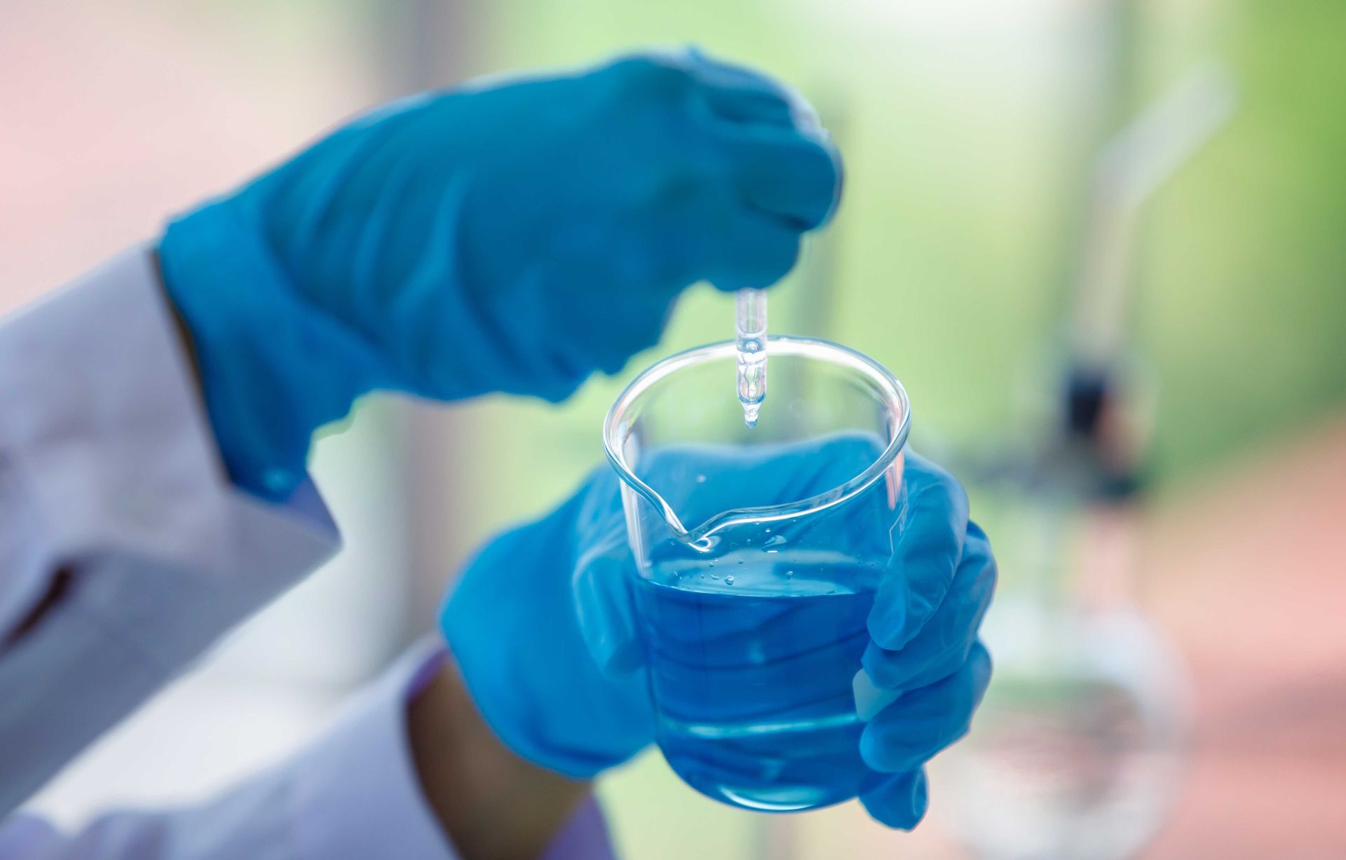 Scientist in blue gloves holding beaker with blue liquid, using a dropper.