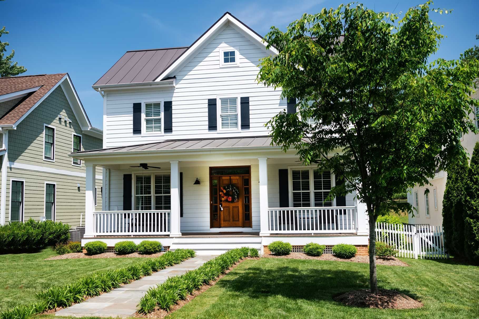 White two-story house with porch, black shutters, brown front door, and green lawn on a sunny day.