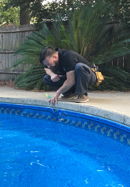Man examining a blue pool, squatting with tool belt. Outdoors, sunny.