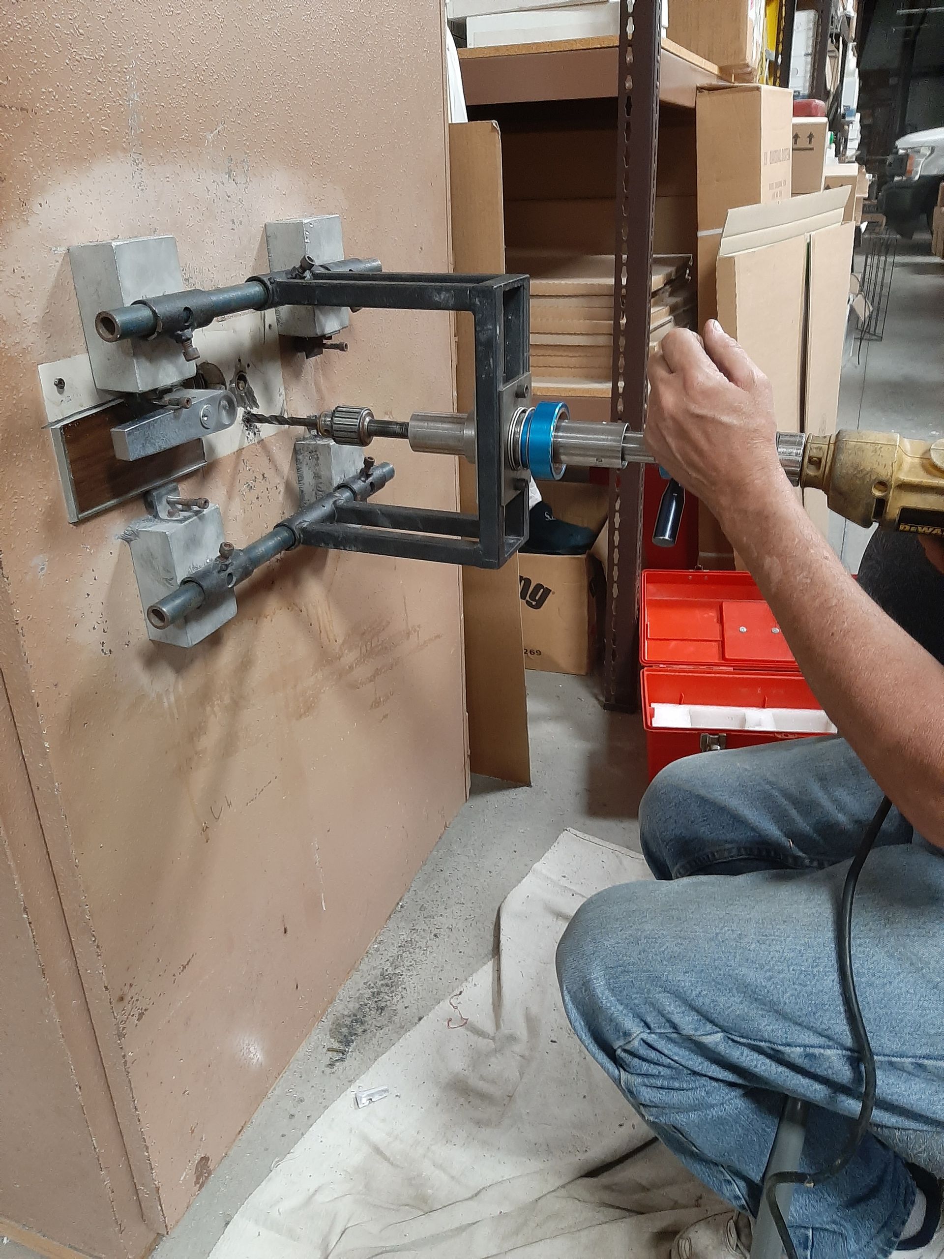 Worker using a drill on a wall-mounted metal bracket in a workshop or storage area.
