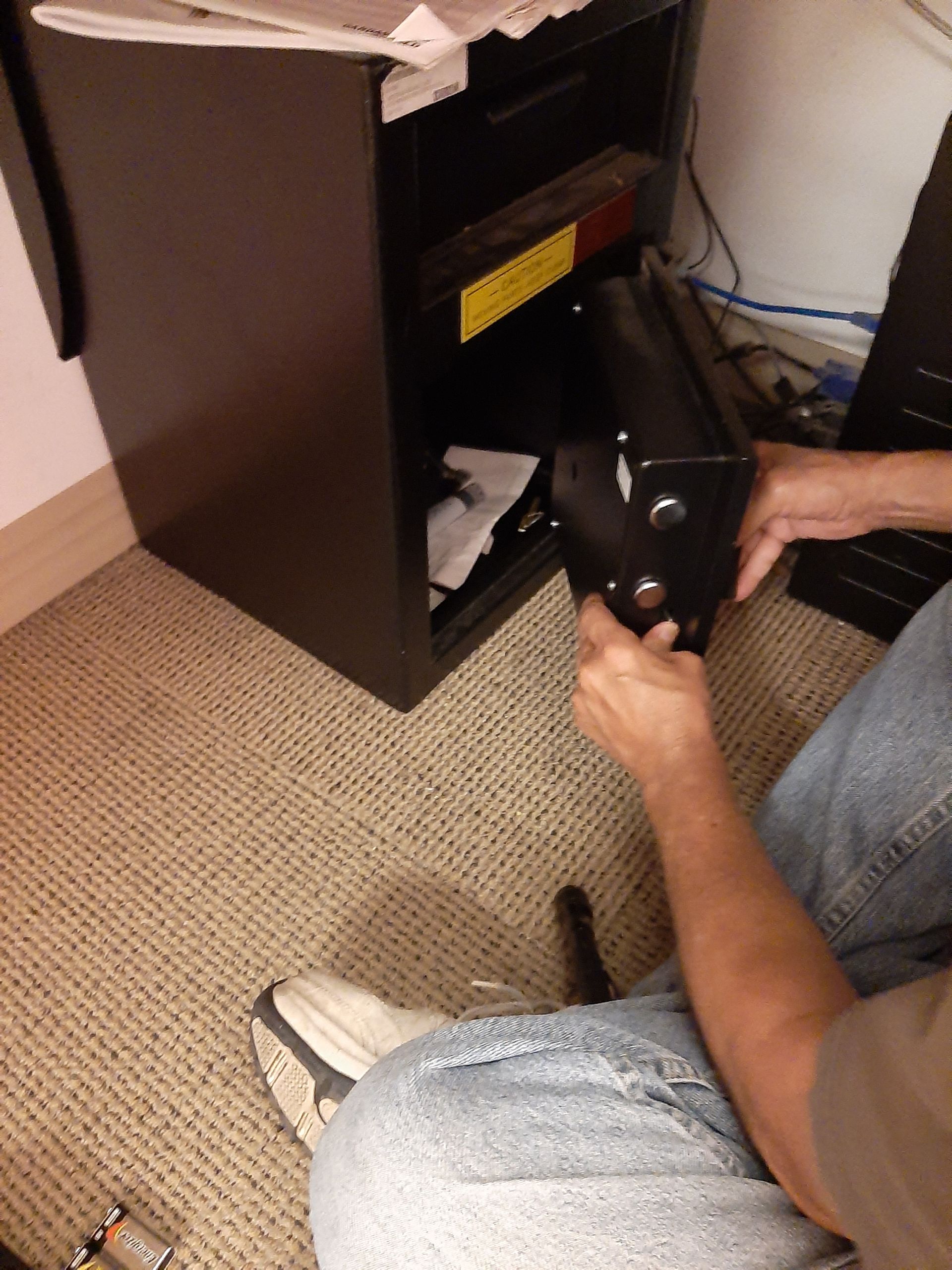 Person kneeling by a black cabinet, reaching into an open lower compartment with a tool.