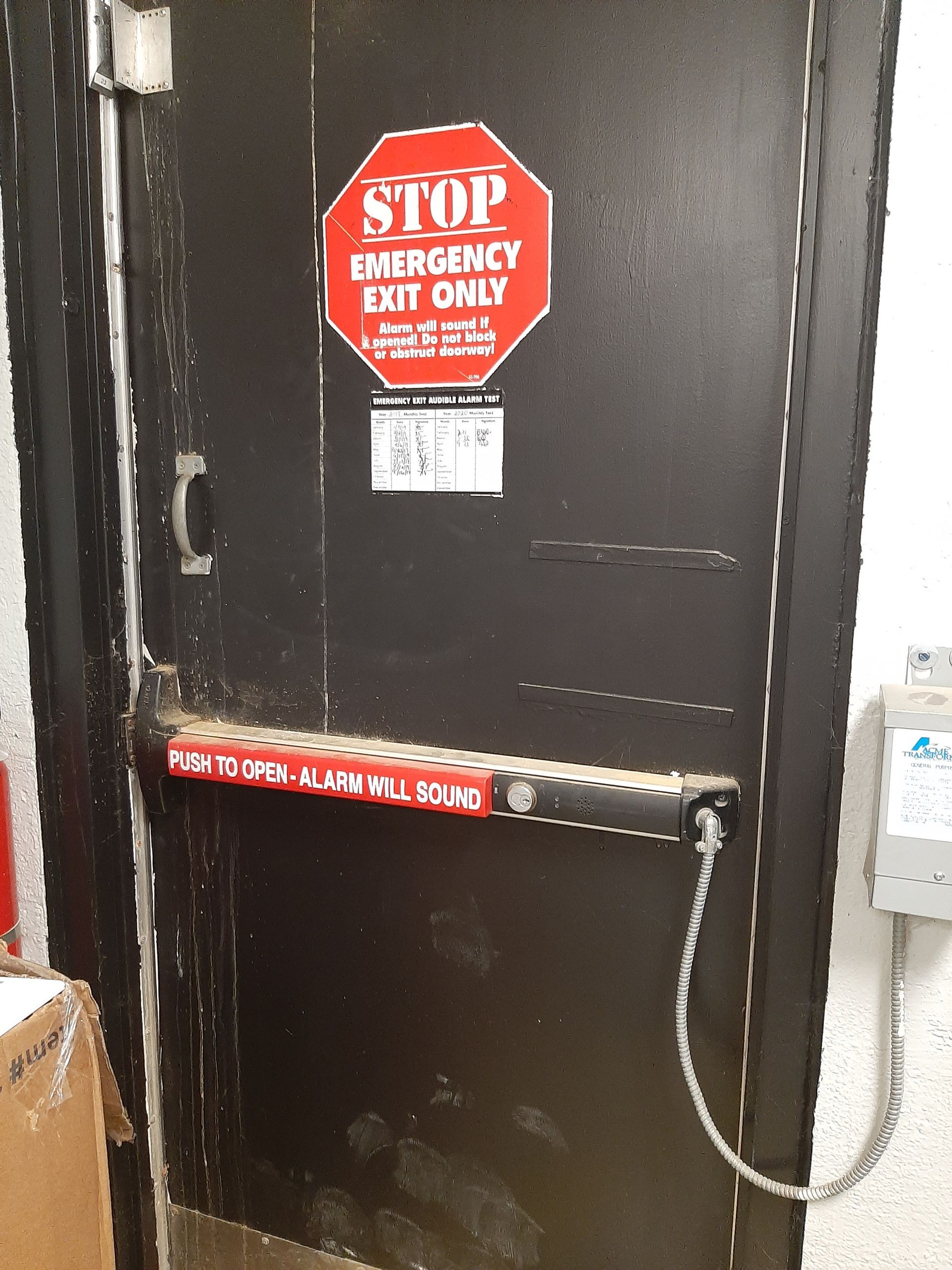 Black emergency exit door with red “STOP” sign and push bar, in an industrial hallway.