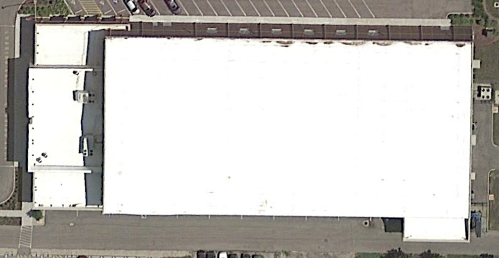 Overhead view of a large rectangular building with a flat white roof, parking areas, and surrounding pavement.