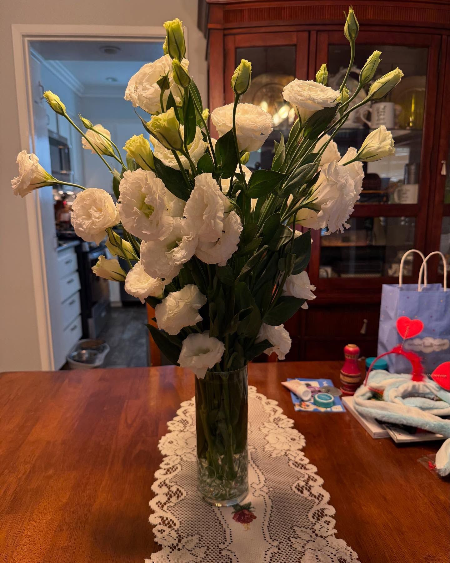 White flowers in a glass vase on a wooden table with a lace runner; a kitchen is in the background. White flowers in a glass vase on a wooden table with a lace runner; a kitchen is in the background.
