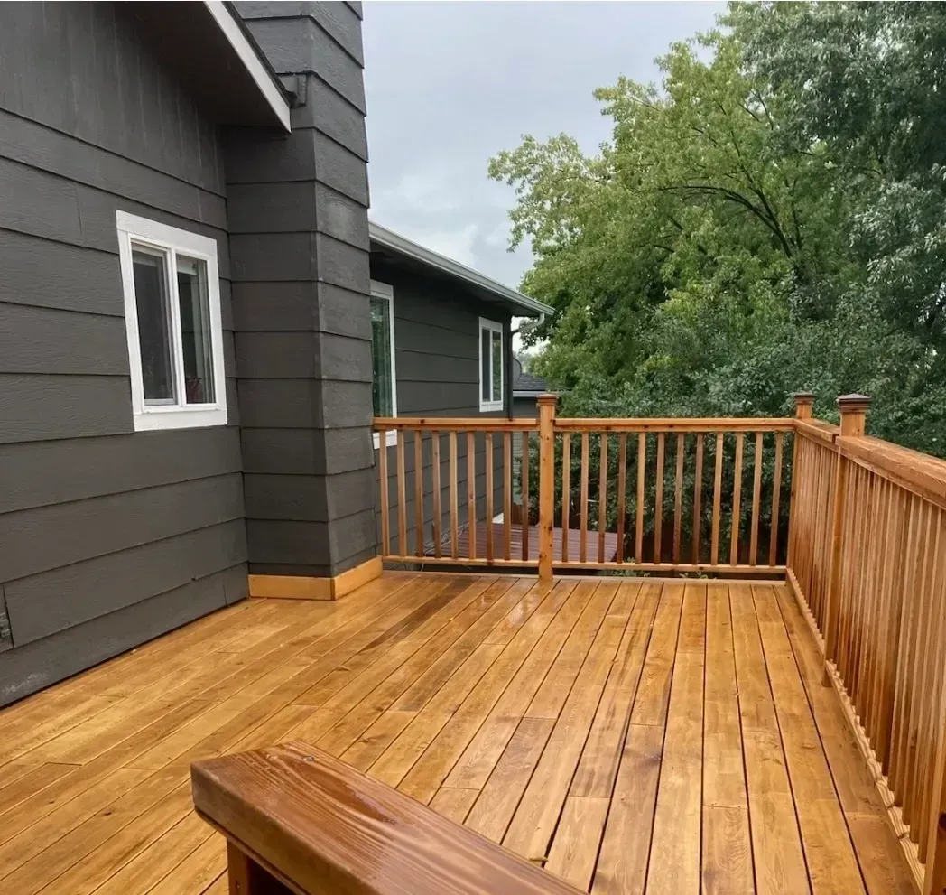 Wooden deck with a wooden railing attached to a dark gray house; green trees are in the background.