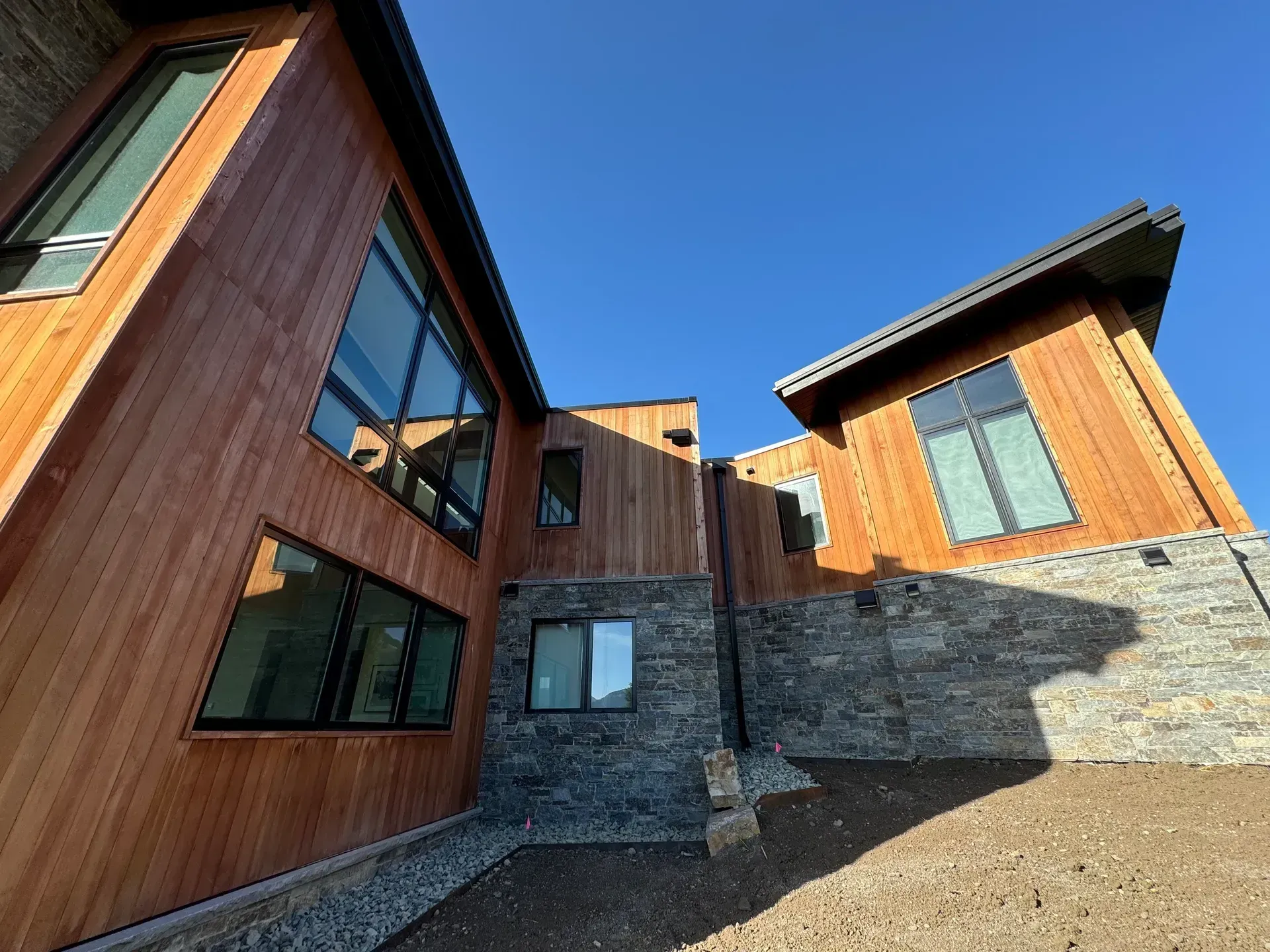 Modern house with wood siding, stone base, and large windows against a clear blue sky.