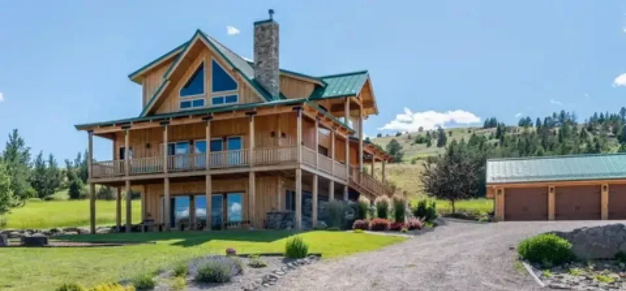 Wooden cabin-style house with green roof and attached garage on a gravel driveway, against a backdrop of hills and blue sky.