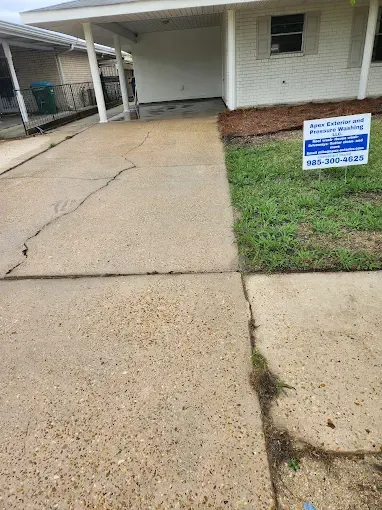 A concrete driveway with cracks leads to a white brick house with a carport. A lawn with a business sign borders the driveway.