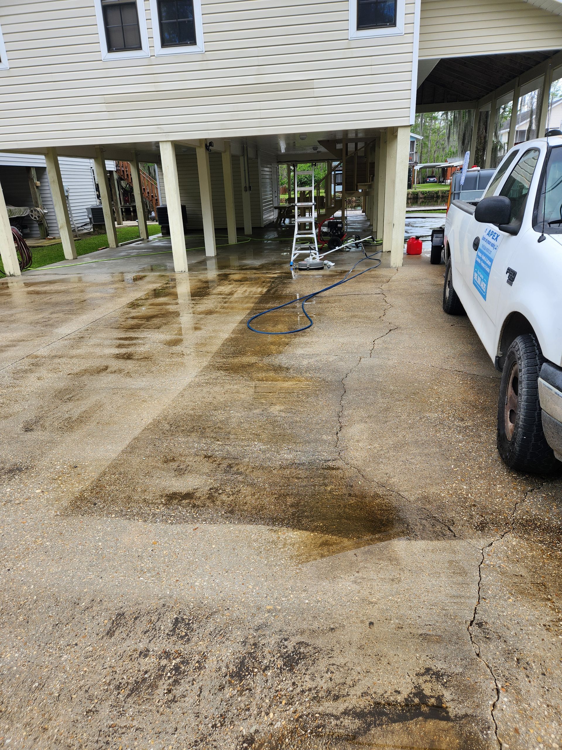 Wet driveway with a house elevated on pillars; a white truck is parked. A ladder and equipment are visible.