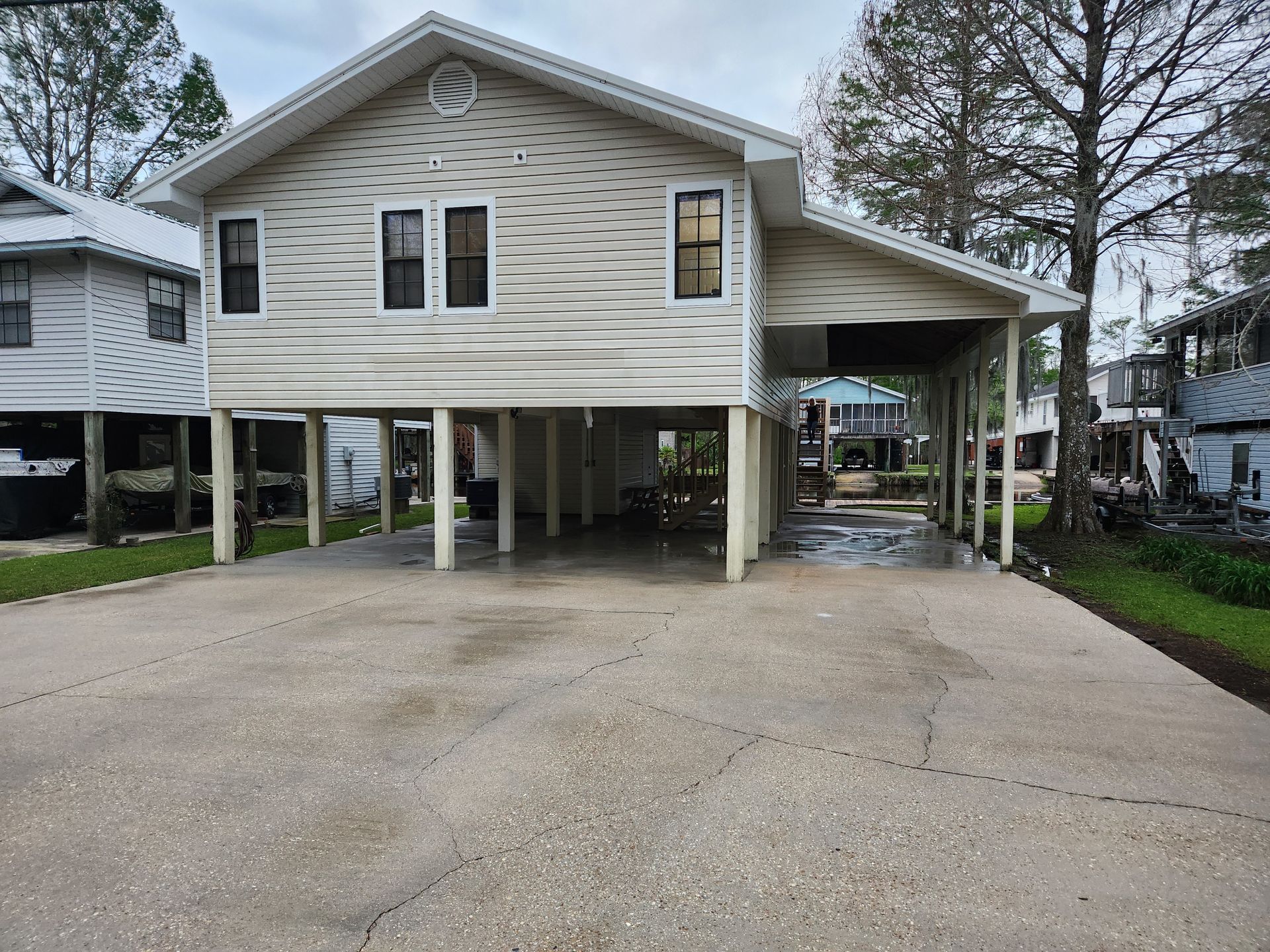 A two-story house on stilts with a concrete driveway and carport. The house is light-colored with a covered parking area on the right side.