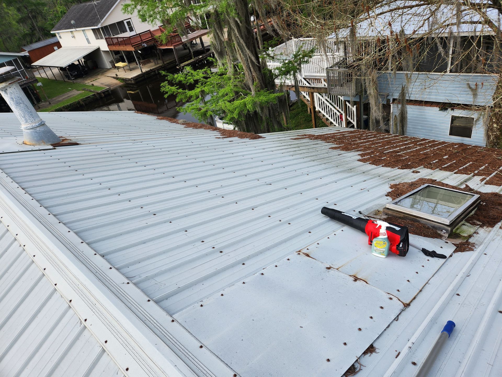 A metal roof with a leaf blower and skylight. The roof is covered in debris. A building and trees are in the background.
