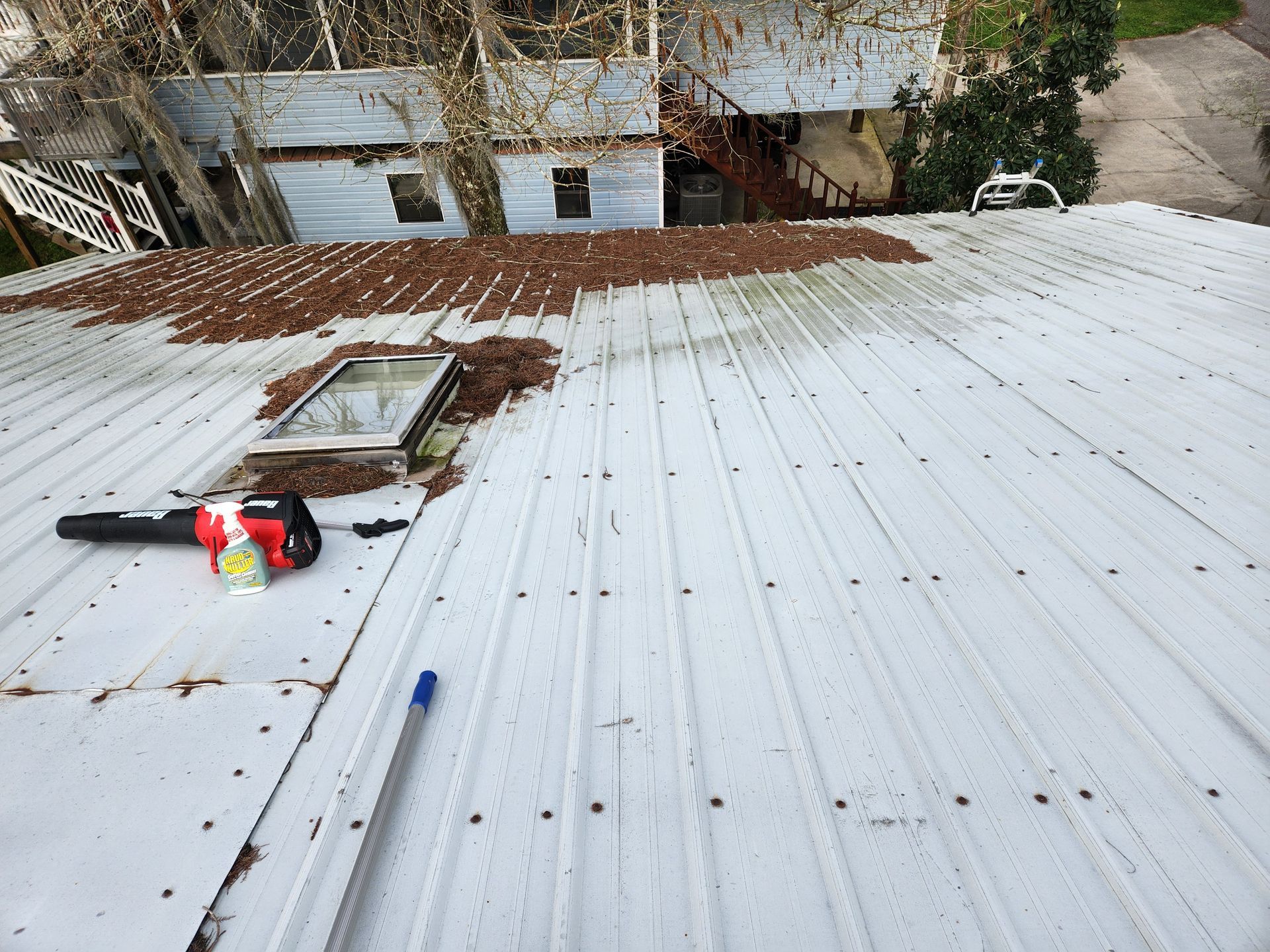 A light gray metal roof with a skylight and debris is shown from an elevated perspective, with a blower and cleaning pole visible.