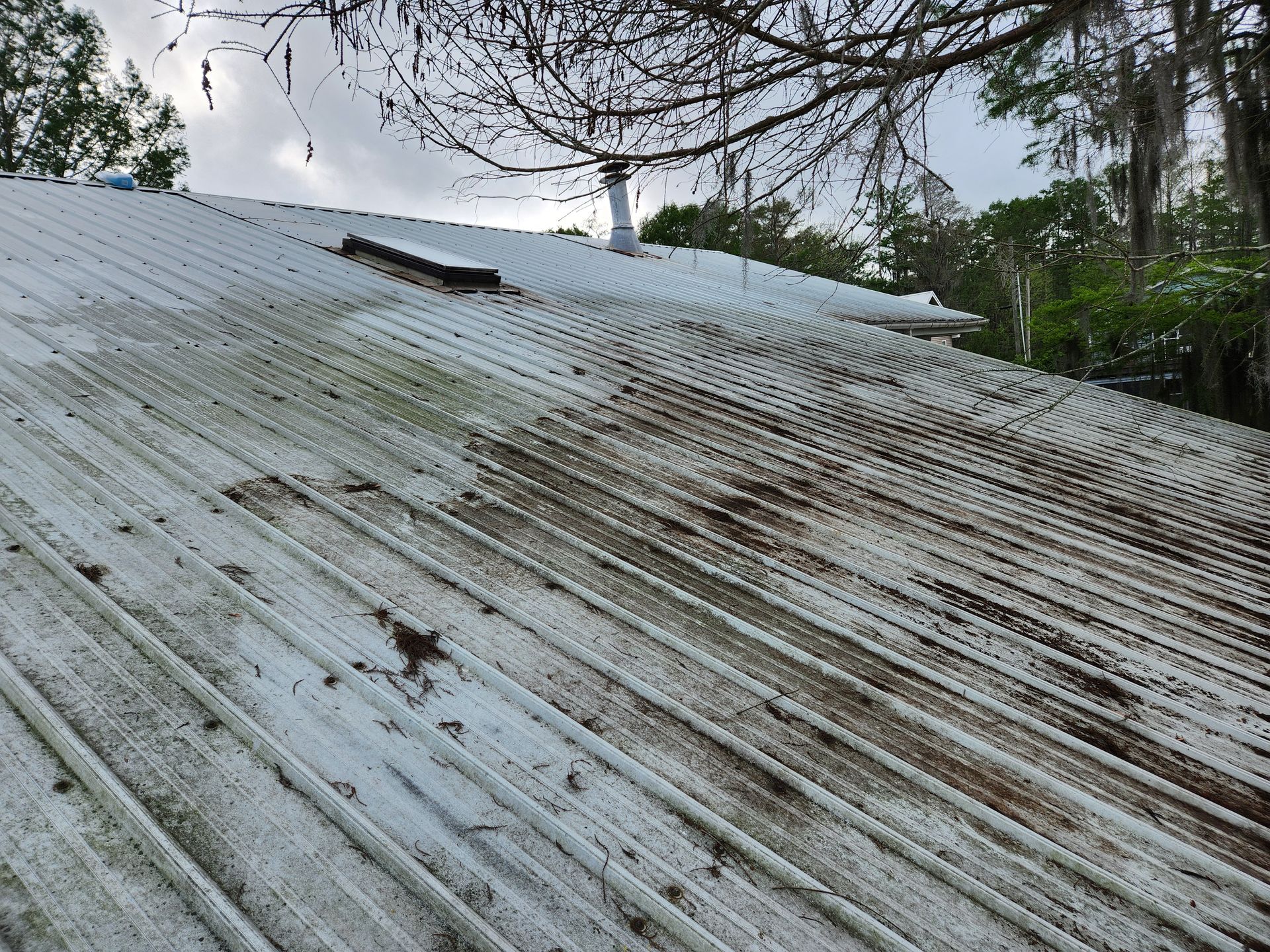 A weathered, white corrugated metal roof with dark streaks of algae, chimney, and a cloudy sky.