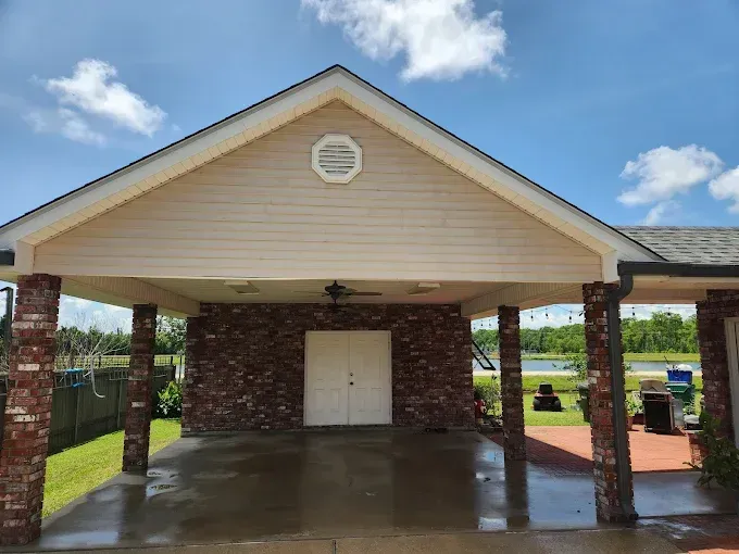 A covered outdoor pavilion with red brick columns and a tan roof, facing a body of water under a blue sky.