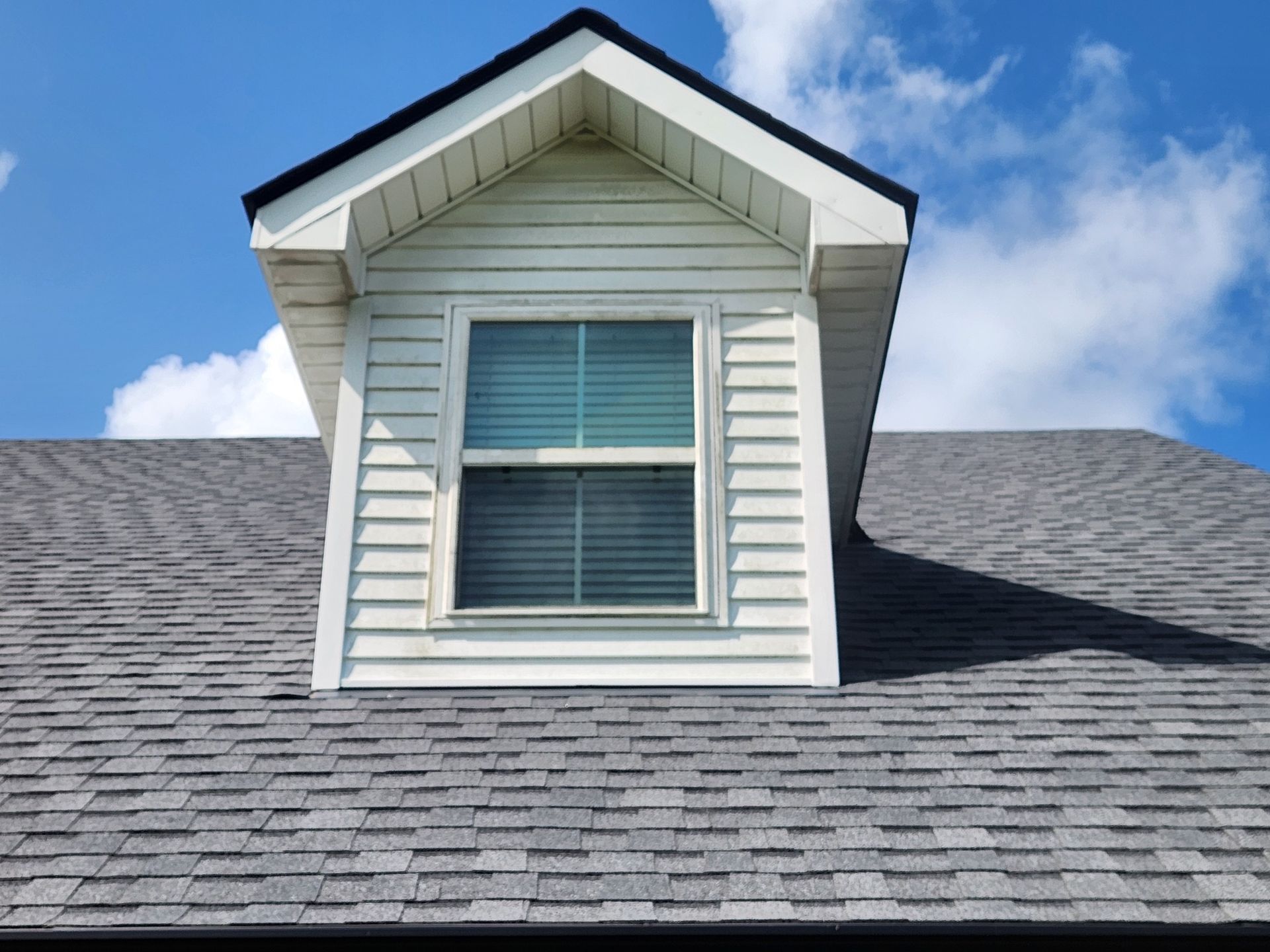 Dormer window with white siding on a gray shingled roof against a blue sky with clouds.