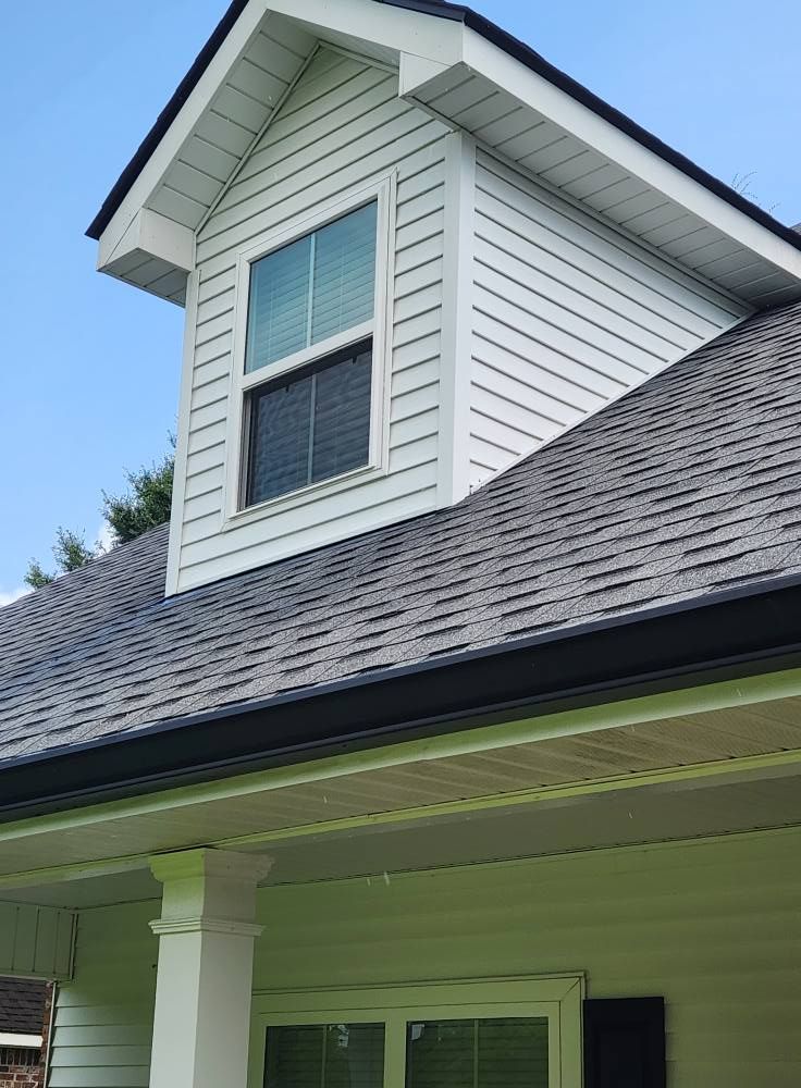 Dormer with a white exterior and black roof. A window is centered, surrounded by white siding. The scene is outdoors.