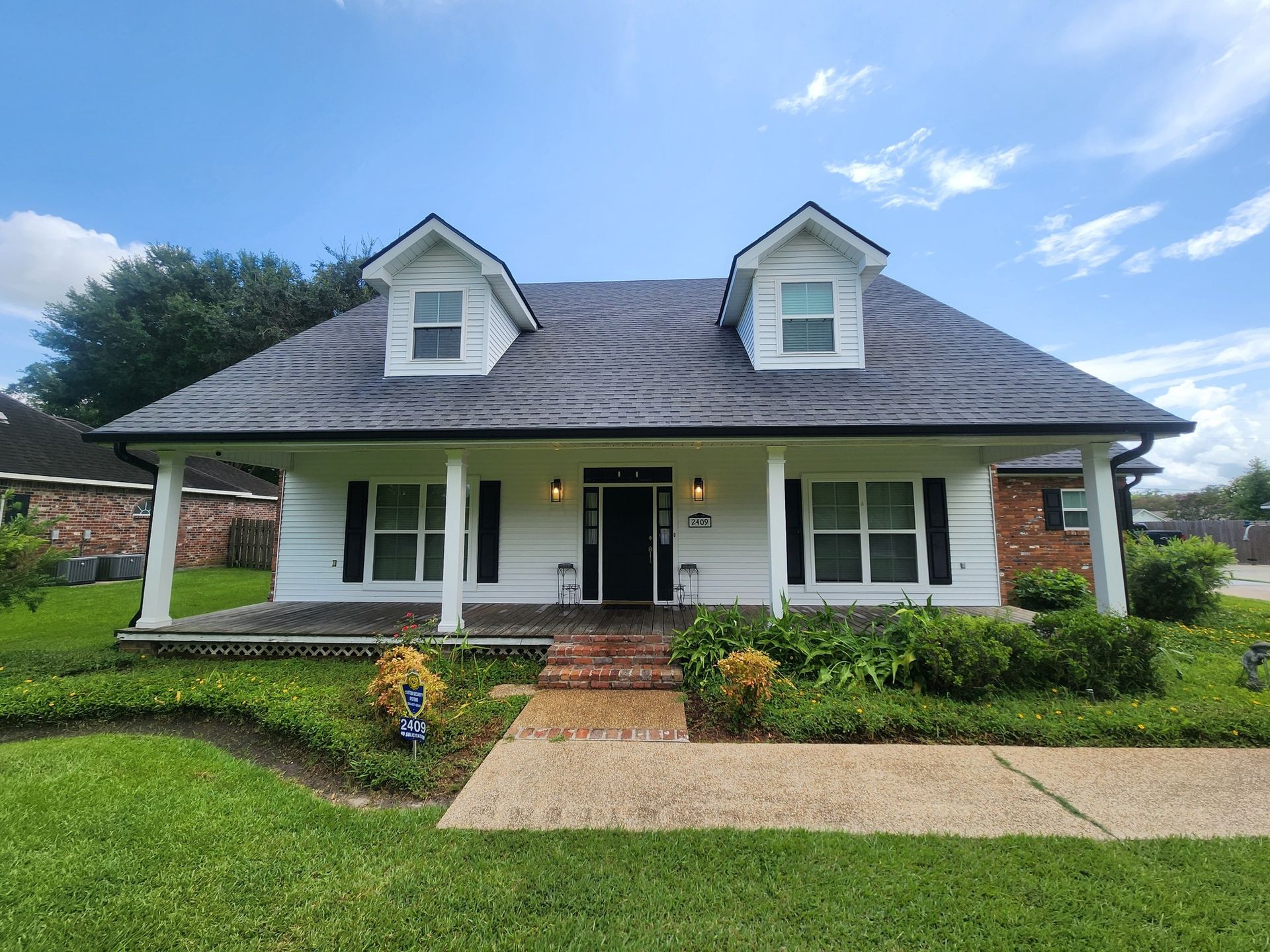 White house with black shutters, a front porch, and two dormer windows under a blue sky. A brick pathway leads to the front door.