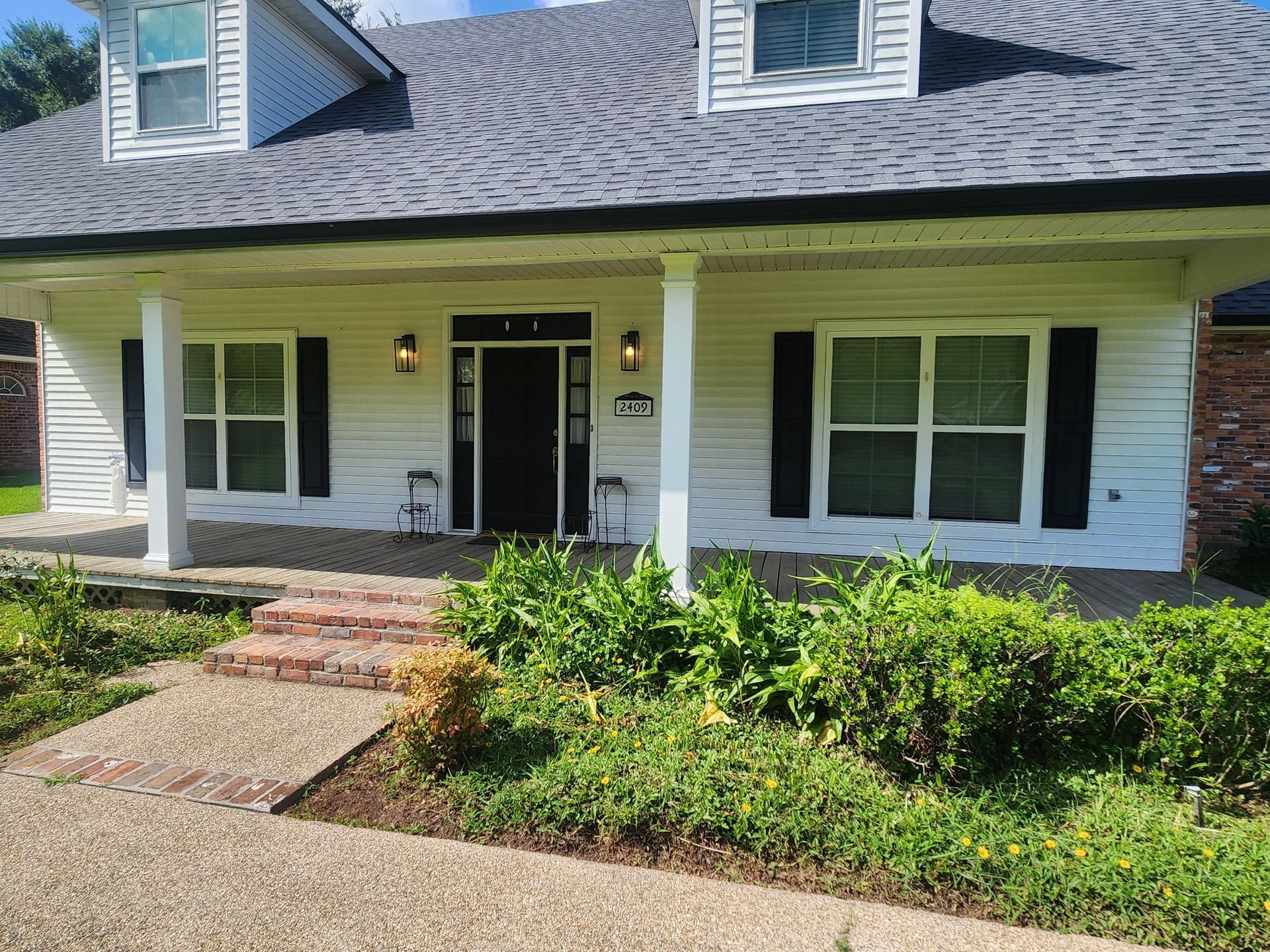 White house with black shutters, columns, and a dark front door. A stone path leads to the entrance, surrounded by green plants.