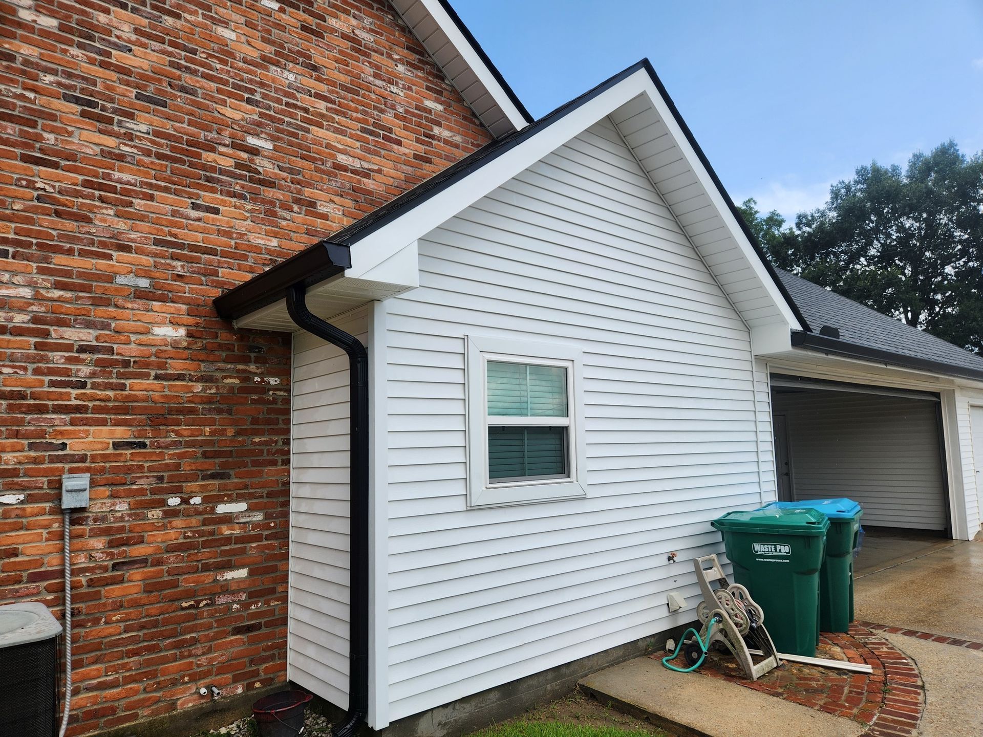 Exterior of a house: Brick wall, white siding, black gutters, a window, and garage doors. Two green trash cans are outside.